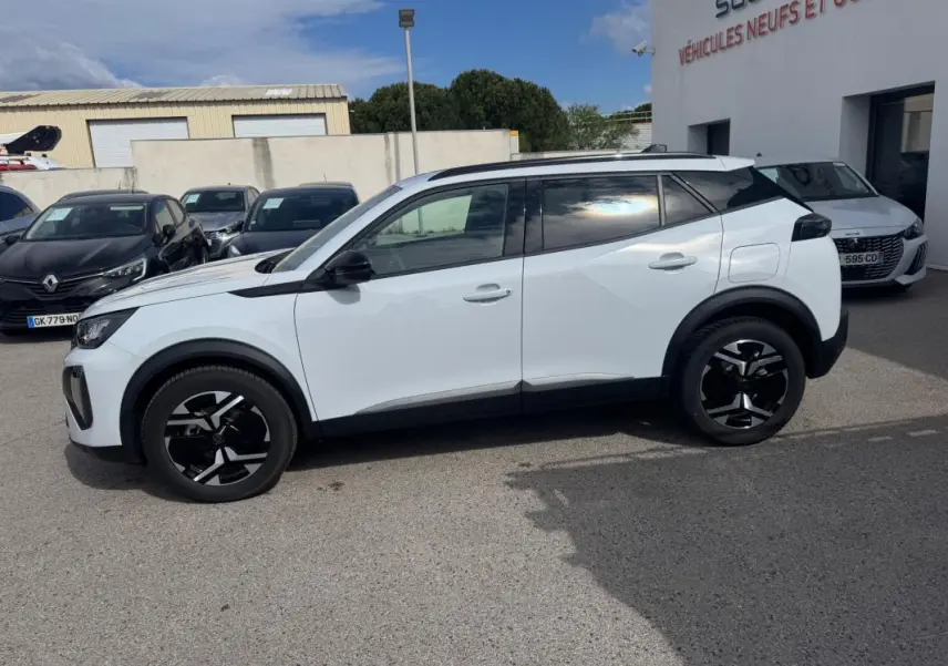 Vue de profil côté gauche d’un Peugeot 2008 blanc 2024 avec jantes alliage noires et barres de toit visibles.