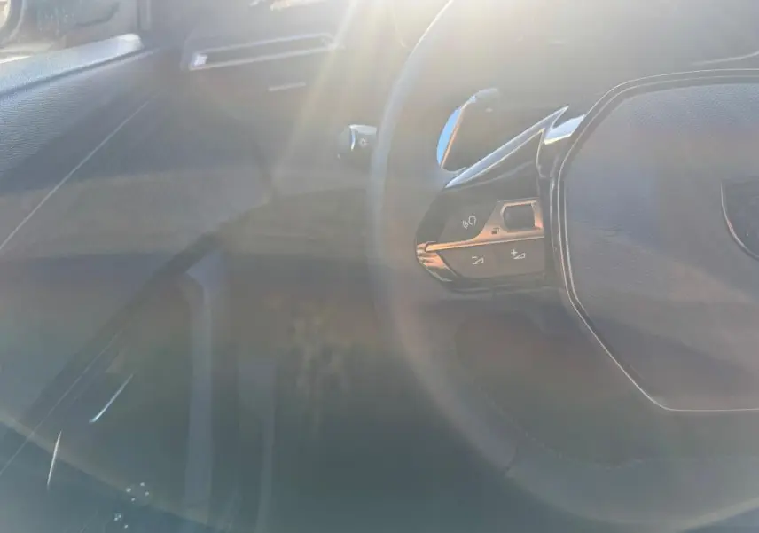 Vue partielle du volant noir du Peugeot 2008 Hybrid 136 e-DCS6 Allure 2024 avec commandes intégrées, sous un fort éclairage naturel.