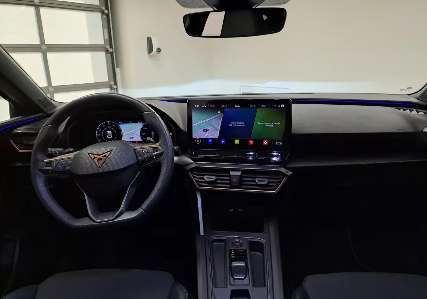 Vue intérieure avant du CUPRA Leon 2024 avec tableau de bord noir, écran tactile central et volant multifonction.