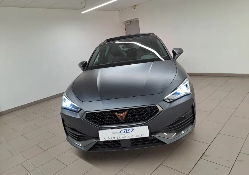 Vue avant d'une CUPRA Leon 2024 gris magnétique mat avec toit ouvrant panoramique et phares allumés en intérieur.