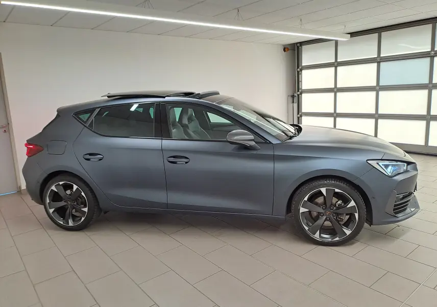 Vue de profil côté gauche d'une CUPRA Leon 2024 gris magnétique mat avec toit ouvrant panoramique en intérieur.