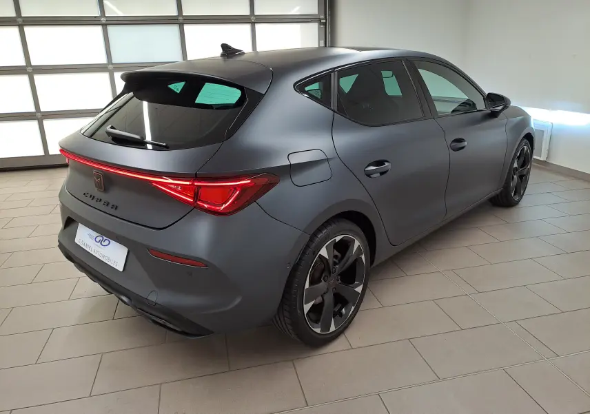 Vue 3/4 arrière droite d'une CUPRA Leon 2024 gris magnétique mat avec toit ouvrant panoramique et feux arrière allumés.