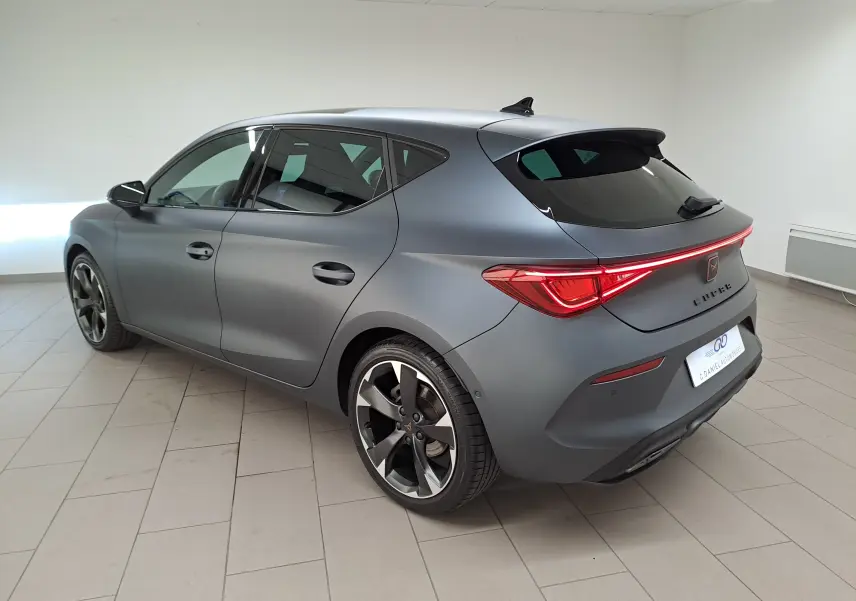 Vue 3/4 arrière droite d'une CUPRA Leon 2024 gris magnétique mat avec toit ouvrant panoramique et feux arrière allumés.