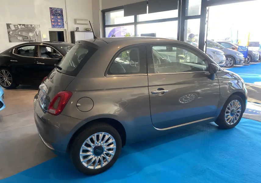 Vue 3/4 arrière droite d'une FIAT 500 Lounge gris foncé stationnée en intérieur sur sol bleu.