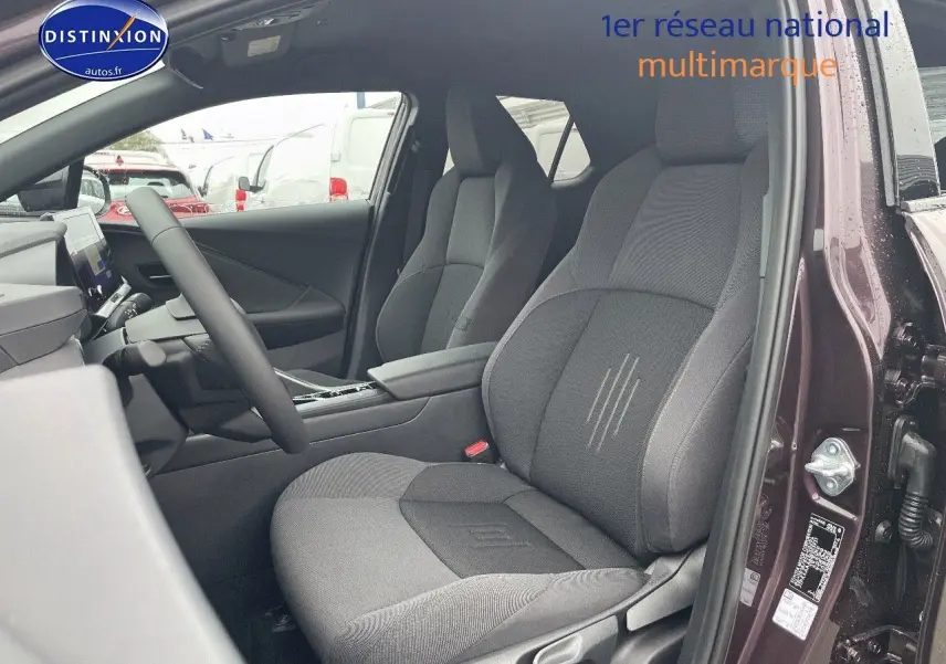 Vue intérieure côté conducteur du Toyota C-HR 1.8 Hybrid 2024 en améthyste métal, sièges tissu gris et console centrale visible.