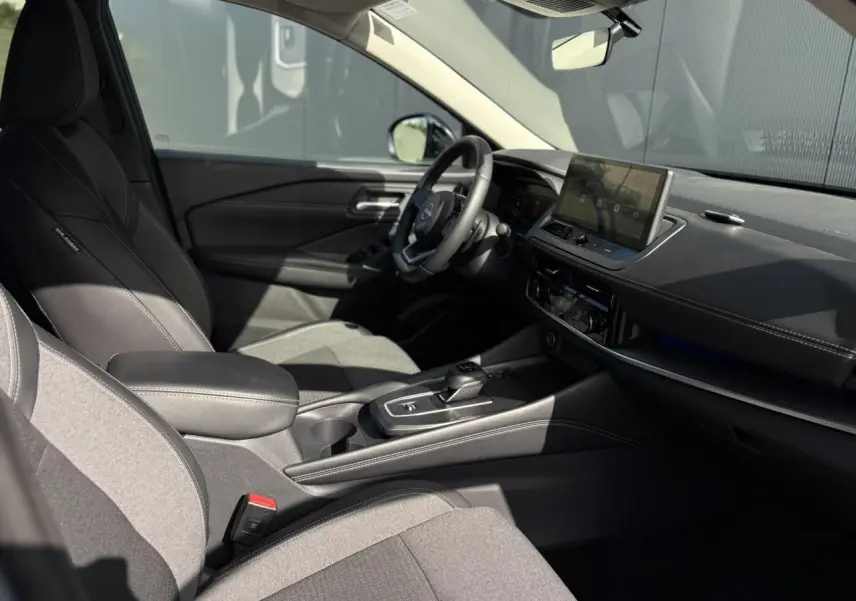Intérieur du Nissan Qashqai 2025 vu côté conducteur, sièges gris, écran tactile et volant cuir noir.