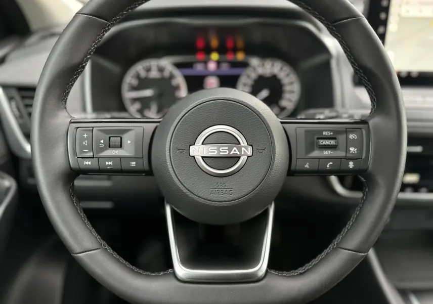 Vue rapprochée du volant en cuir noir du Nissan Qashqai 2024 avec commandes intégrées et tableau de bord flou en arrière-plan.