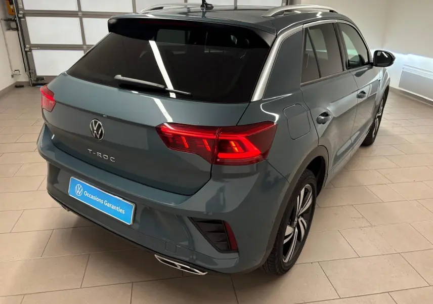 Vue 3/4 arrière droite du Volkswagen T-Roc 2024 bleu pétrole avec feux arrière LED et finition R-Line en intérieur showroom.