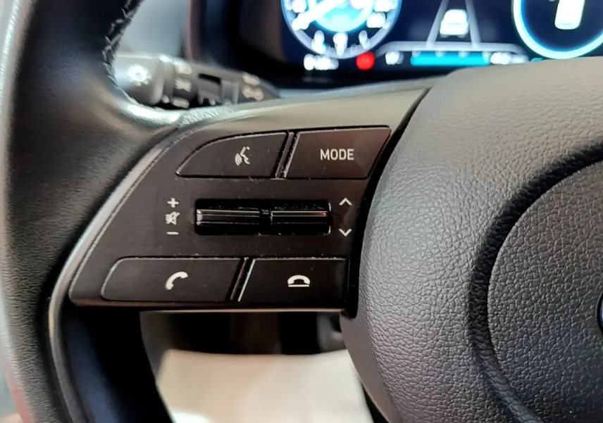 Gros plan sur les commandes multifonctions en cuir du volant gauche de la Hyundai i20 gris clair, tableau de bord numérique en arrière-plan.