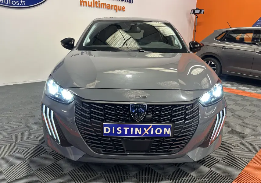Vue frontale d'une Peugeot 208 gris Selenium avec phares allumés et calandre noire distinctive en showroom.