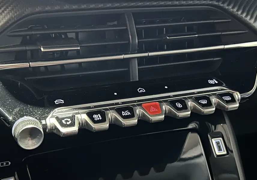 Gros plan sur la rangée de boutons chromés et l'écran tactile noir du tableau de bord intérieur du Peugeot 208 gris Selenium.