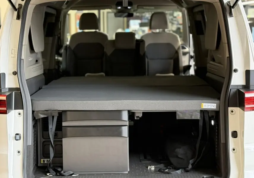 Vue arrière intérieure du Volkswagen California T7 blanc Candy avec couchette déployée et rangements sous le lit.