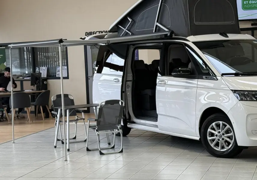 Vue de côté droit d’un Volkswagen California T7 blanc Candy avec toit relevable noir et auvent déployé, porte coulissante ouverte.