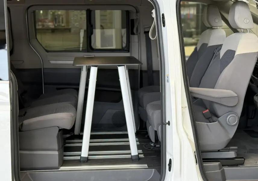 Vue latérale droite du Volkswagen California T7 blanc Candy, porte coulissante ouverte sur l'intérieur avec table repliable entre les sièges.