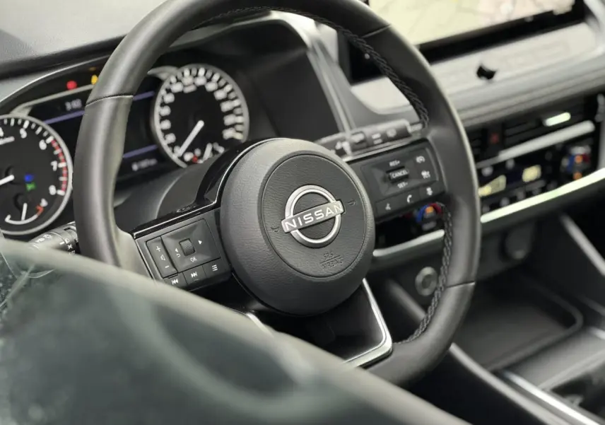 Vue rapprochée du volant noir du Nissan Qashqai 2025 avec commandes intégrées et tableau de bord digital flou.