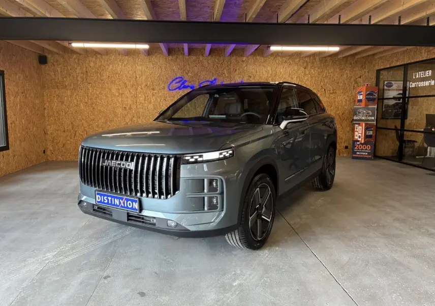 SUV JAECOO J7 gris olive en 3/4 avant droit, avec calandre verticale noire et toit noir dans un garage.