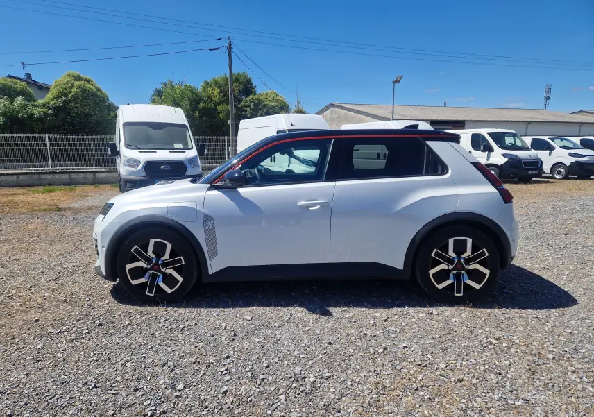 Profil droit de la Renault R5 E-TECH 2025 blanc nacré avec toit noir et jantes stylisées sur un parking extérieur.