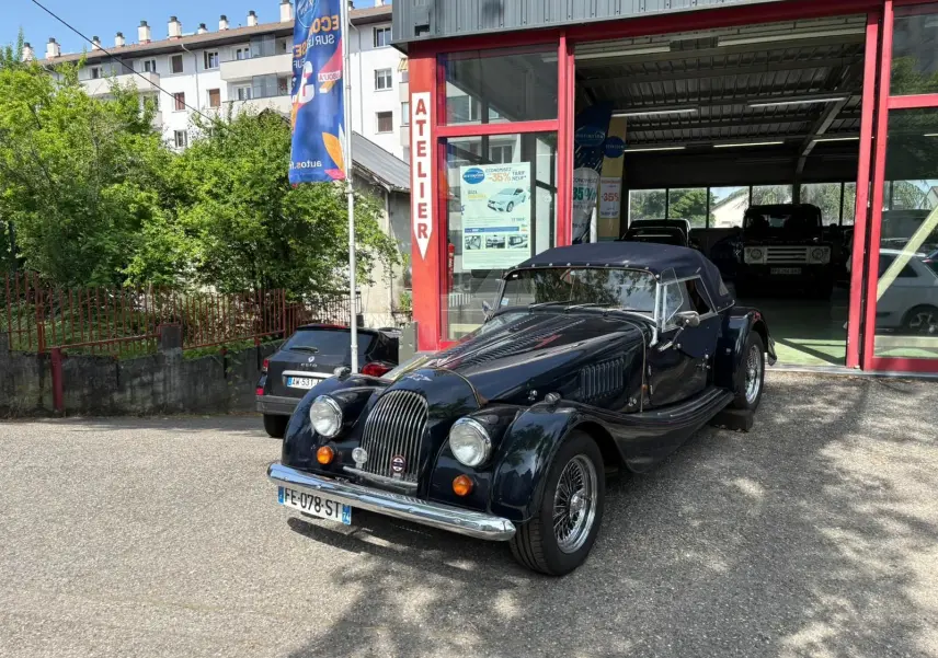 Morgan Plus 4 bleu foncé vue 3/4 avant droit, capote noire et calandre verticale chromée devant un atelier.