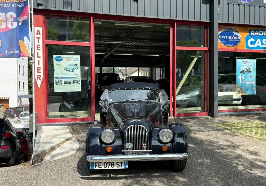 Morgan Plus 4 bleu foncé vue de face, capote noire et calandre chromée devant un atelier automobile.