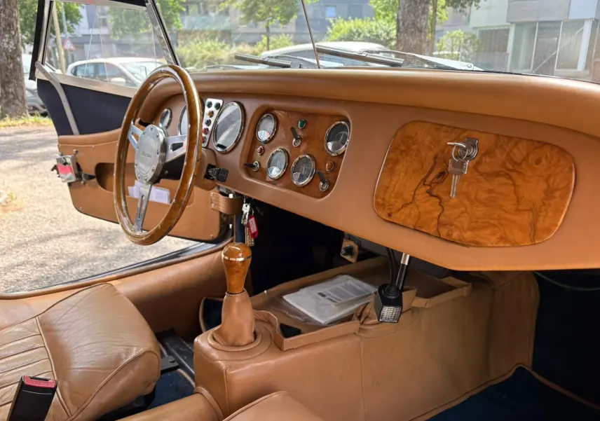 Intérieur beige de la Morgan Plus 4 1995, vue côté conducteur sur tableau de bord en bois et volant bois.