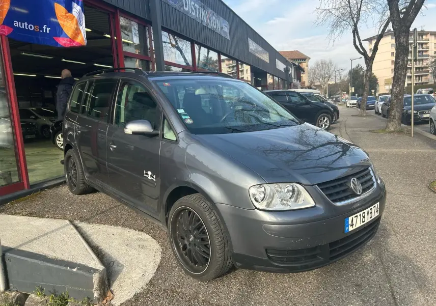 Volkswagen Touran gris vue 3/4 avant gauche avec jantes noires et sticker sur la porte côté gauche.