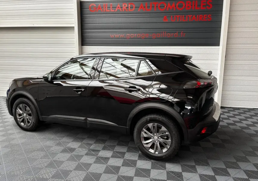 Peugeot 2008 noir vue de profil côté gauche dans un garage avec sol à damier et panneau publicitaire en arrière-plan.