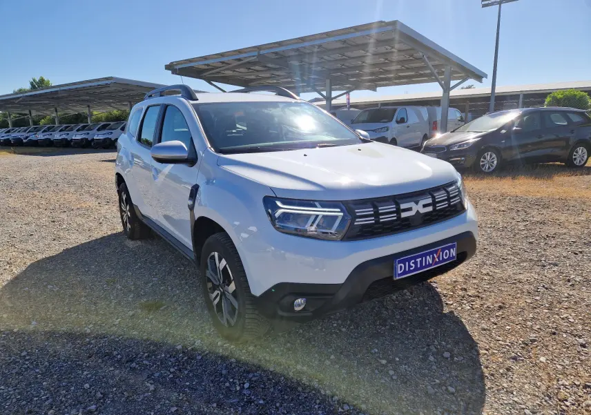 Vue 3/4 avant d'un Dacia Duster blanc 2024 avec jantes alu 17 pouces, sur un parking extérieur ensoleillé.