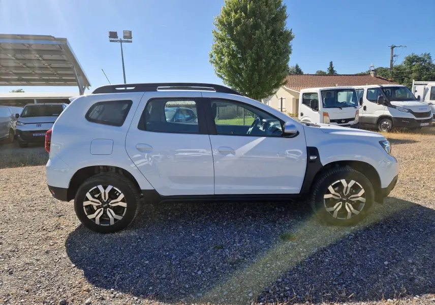 Vue latérale droite d'un Dacia Duster blanc 2024 avec jantes alu 17 pouces et barres de toit noires.