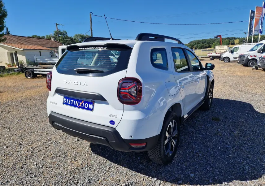 Vue 3/4 arrière droite d'un Dacia Duster blanc 4x4 2024 avec jantes alu 17 pouces sur un terrain gravillonné.