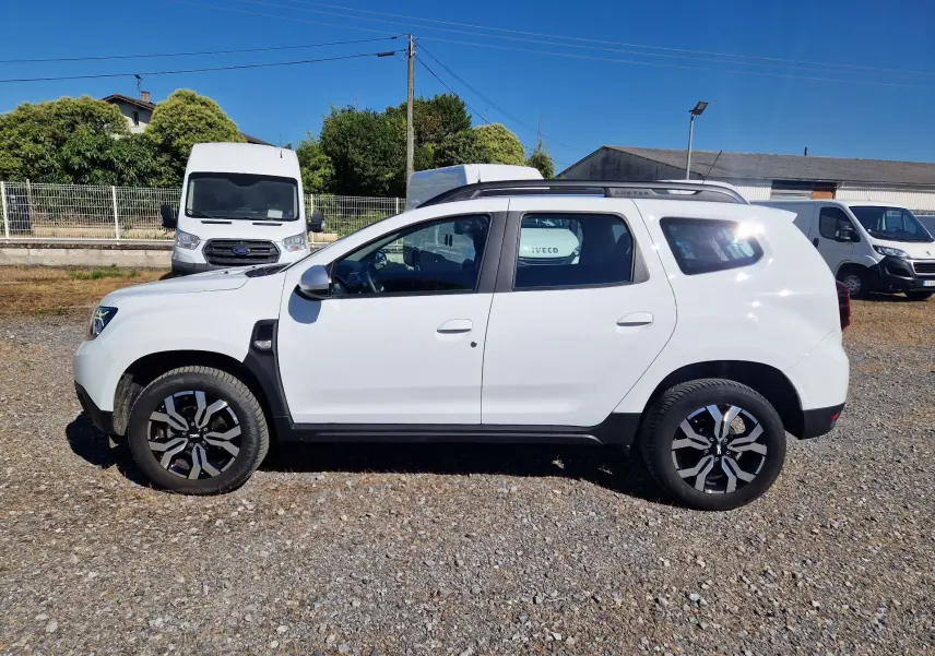 Vue de profil côté gauche d'un Dacia Duster blanc 4x4 2024 avec jantes alu 17 pouces sur un sol gravillonné.
