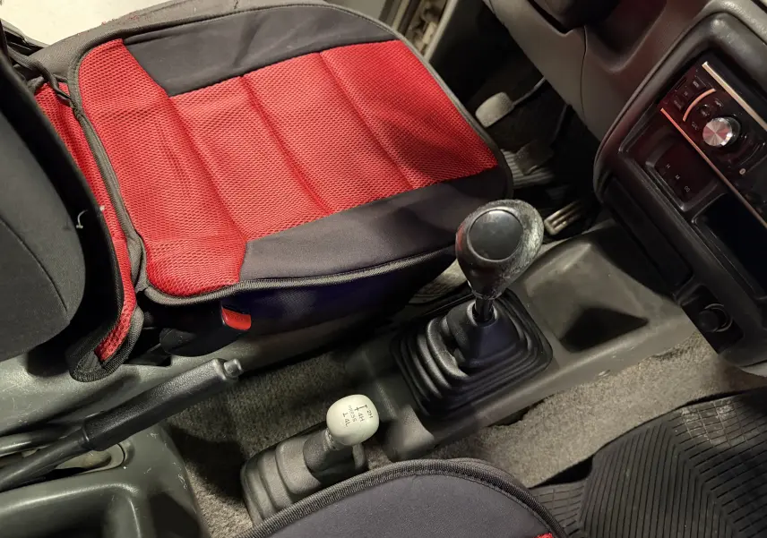 Vue intérieure centrée sur la console centrale d’un Suzuki Jimny Cabriolet gris, montrant levier de vitesse manuel et siège avec coussin rouge.