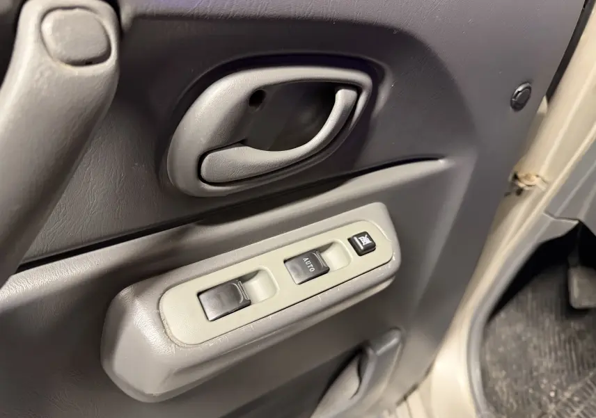 Gros plan sur la poignée intérieure et les commandes des vitres électriques côté gauche d’un Suzuki Jimny Cabriolet gris 2005.