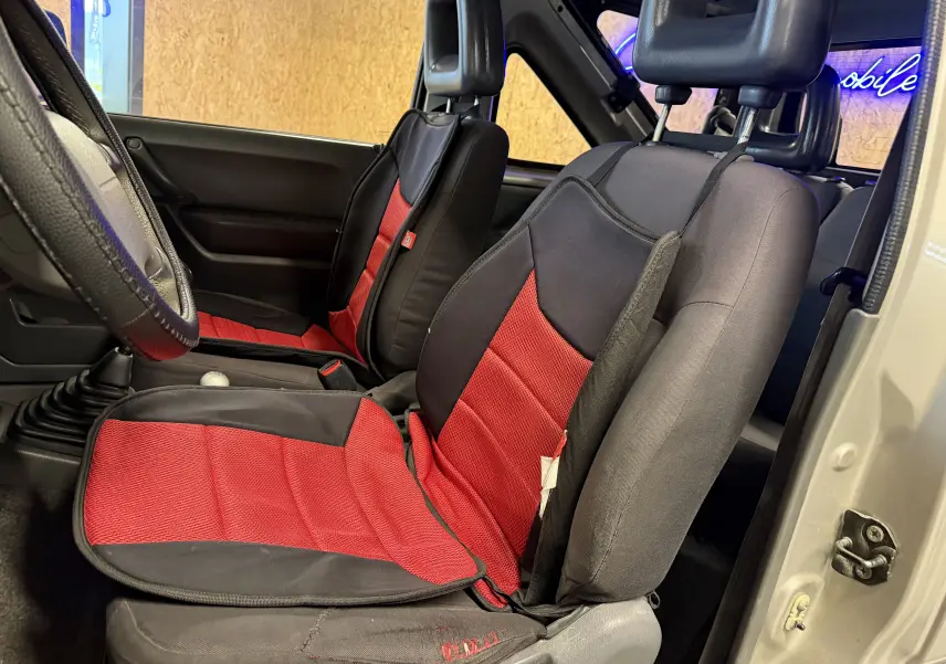 Vue intérieure côté conducteur du Suzuki Jimny Cabriolet gris 2005, sièges avant avec housses rouges et noires.