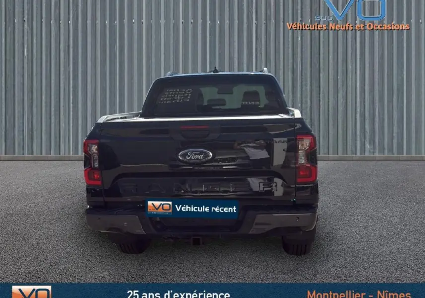 Vue arrière d'un Ford Ranger Wildtrak noir 2025 avec attelage et barres de toit visibles sur fond industriel.