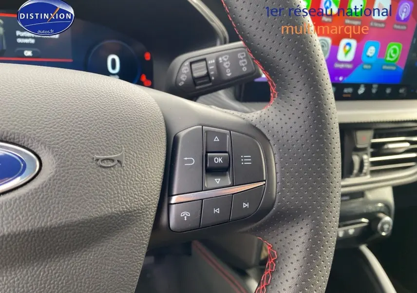 Gros plan sur le volant perforé avec surpiqûres rouges du Ford Kuga 2025, écran tactile et tableau de bord numériques visibles.