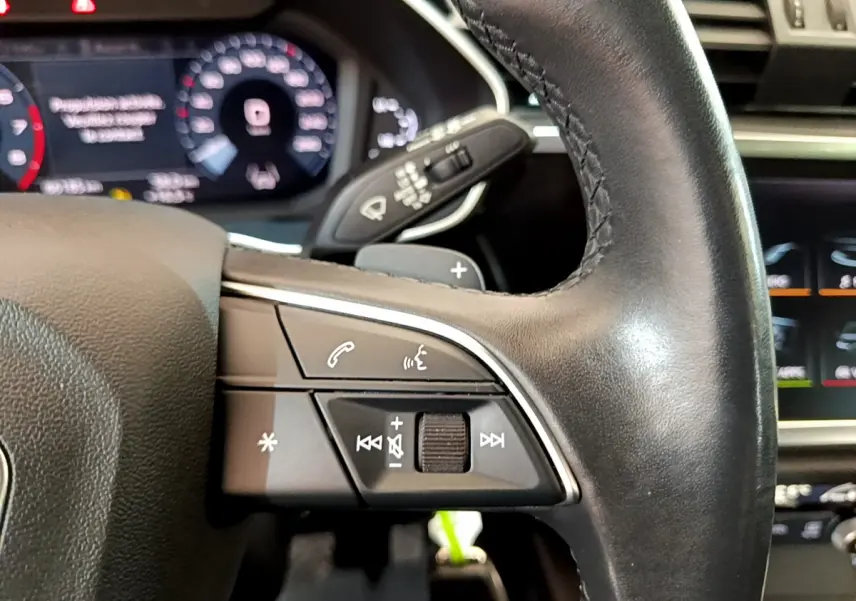 Gros plan sur le volant cuir noir de l'Audi Q3 Sportback 35 TFSI, avec commandes audio et affichage numérique en arrière-plan.