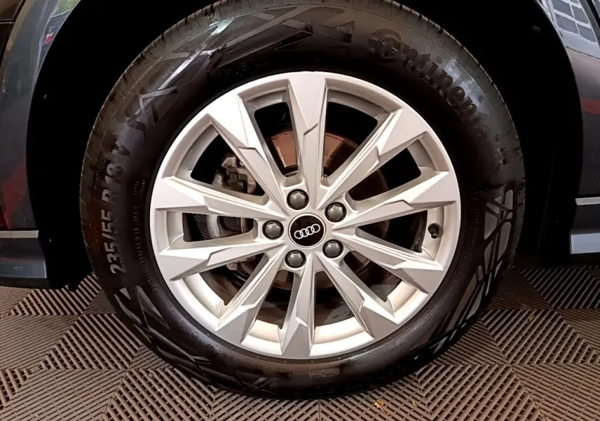 Gros plan sur la roue avant droite avec jante argentée et logo Audi, partie noire de la carrosserie visible en arrière-plan.