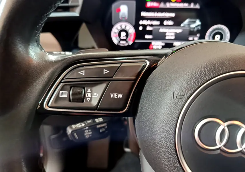 Gros plan sur le volant cuir noir de l'Audi A3 bleu 2023, avec commandes multifonctions et tableau de bord numérique flou.