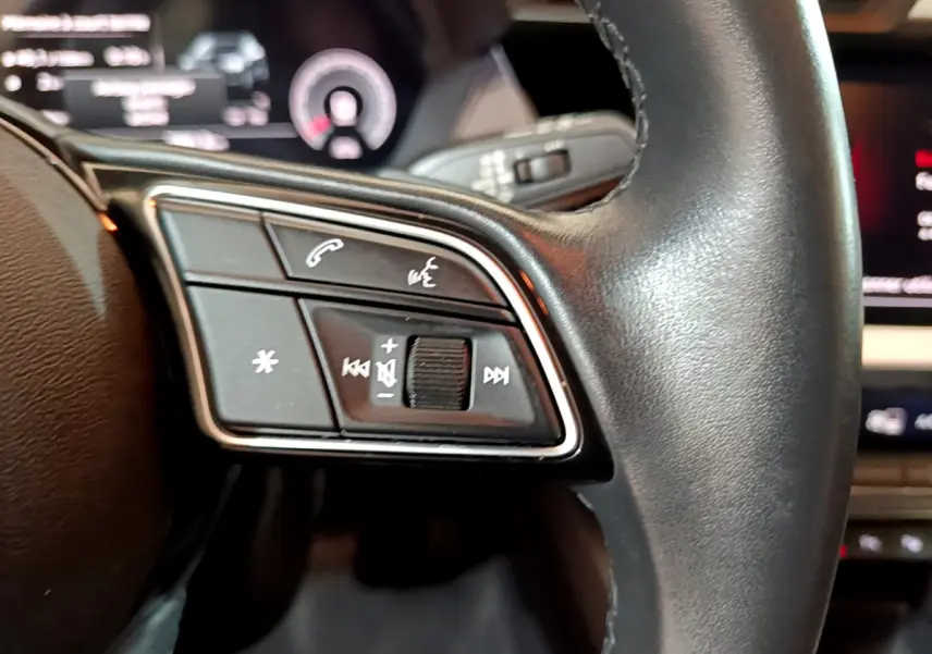 Gros plan sur les commandes gauche du volant cuir de l'Audi A3 35 TFSI bleu, avec affichage numérique en arrière-plan.