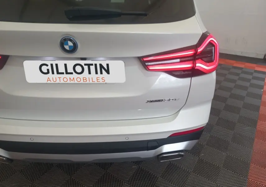 Vue arrière d’un BMW X3 xDrive30e blanc avec feux arrière LED allumés et double sortie d’échappement visible.