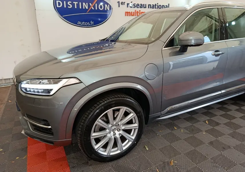Vue 3/4 avant gauche du Volvo XC90 gris clair avec jantes alliage et prise de recharge visible sur l'aile avant.