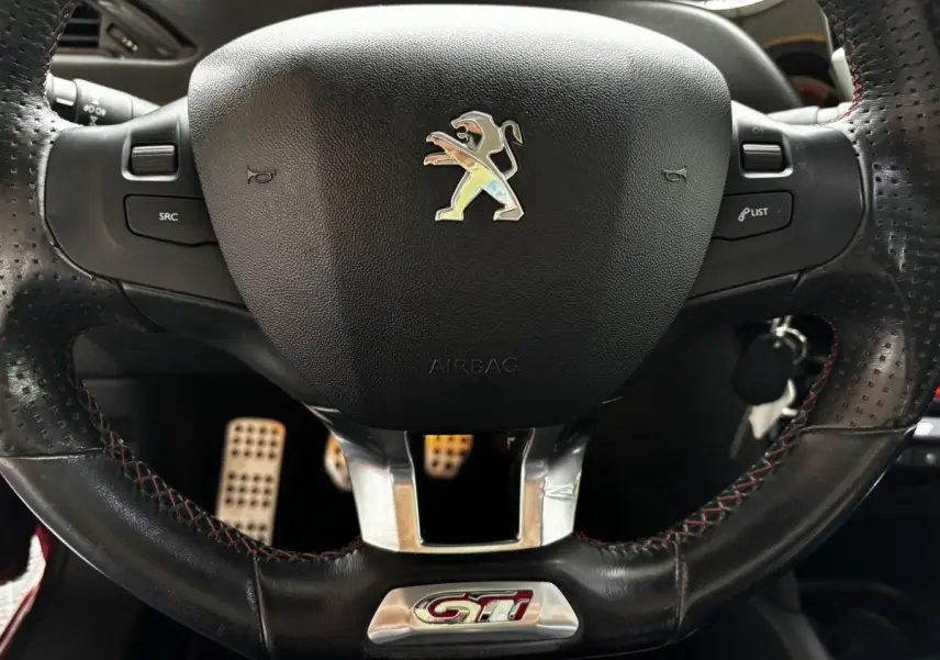 Gros plan sur le volant cuir noir perforé avec logo Peugeot et sigle GTI rouge, pédales sport en arrière-plan.