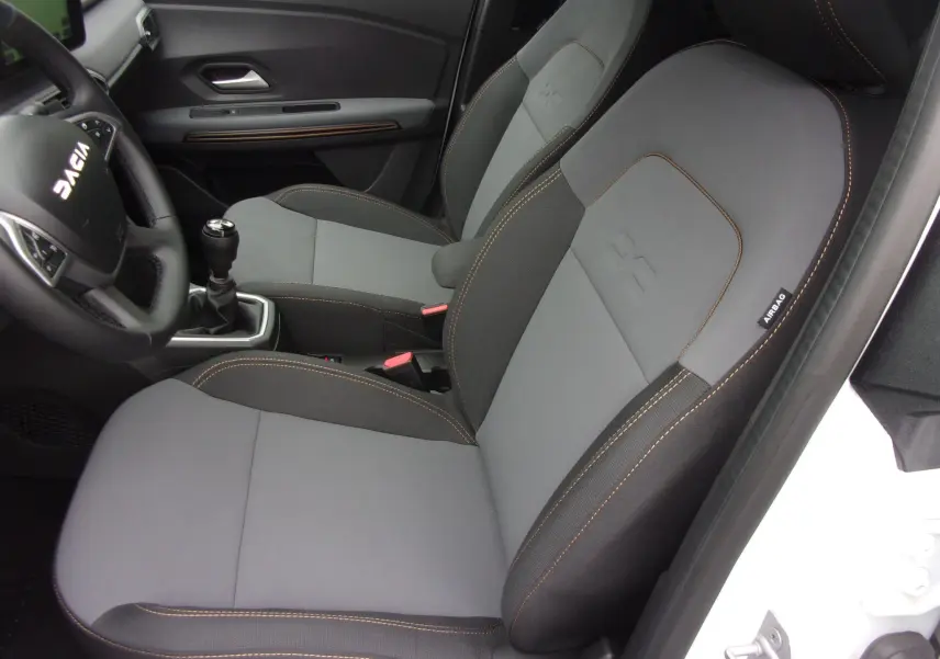 Vue intérieure côté conducteur du Dacia Jogger blanc 2025, sièges gris avec surpiqûres orange et volant marqué DACIA.