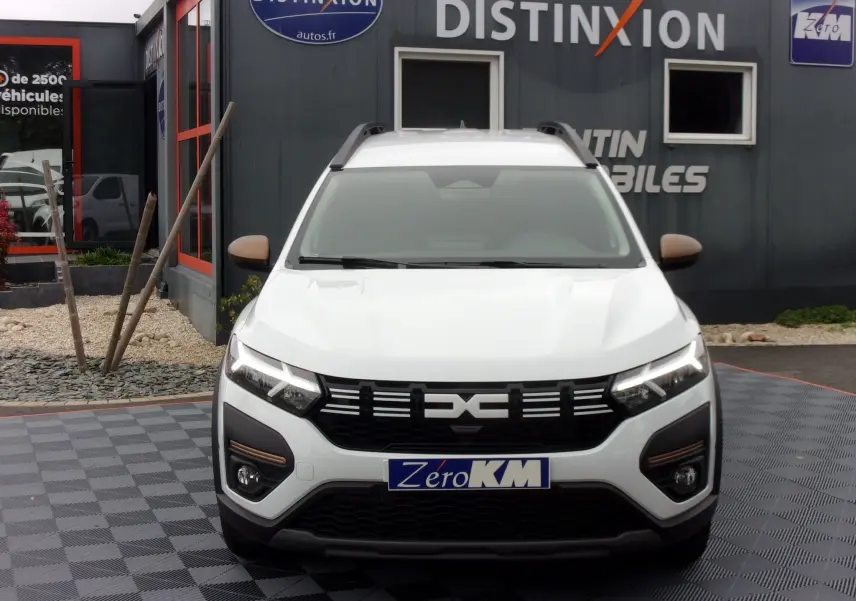 Vue frontale d'un Dacia Jogger blanc 2025 avec calandre noire et phares LED allumés, sur parking de concession.