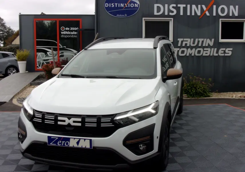 Dacia Jogger blanc en 3/4 avant droit, avec calandre noire et feux LED allumés, stationné devant un concessionnaire.