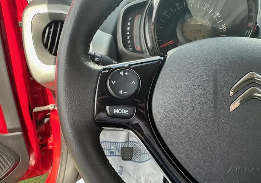 Gros plan sur le volant noir de la Citroën C1 rouge Scarlet, avec commandes audio intégrées et tableau de bord visible.