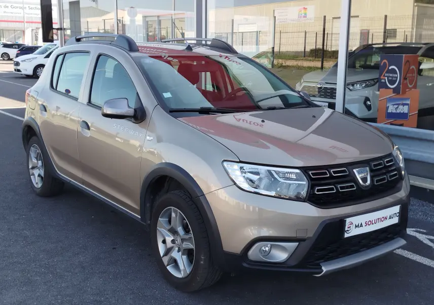 Vue 3/4 avant d'une Dacia Sandero Stepway beige dune avec barres de toit et jantes alliage, stationnée devant un garage.
