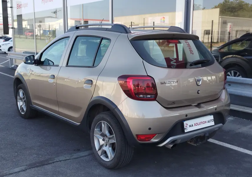 Vue 3/4 arrière droite d'une Dacia Sandero Stepway beige dune avec barres de toit et attelage visible en stationnement.