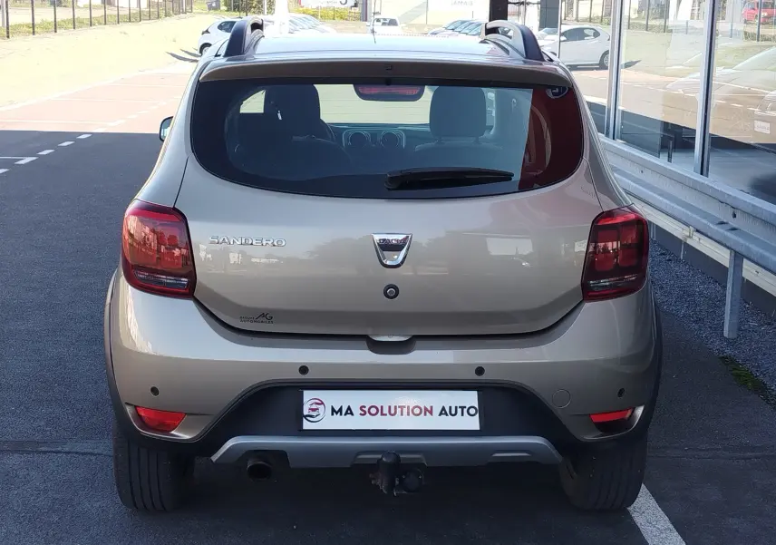 Vue arrière d'une Dacia Sandero Stepway beige dune 2020 avec attelage et feux arrière foncés sur parking.