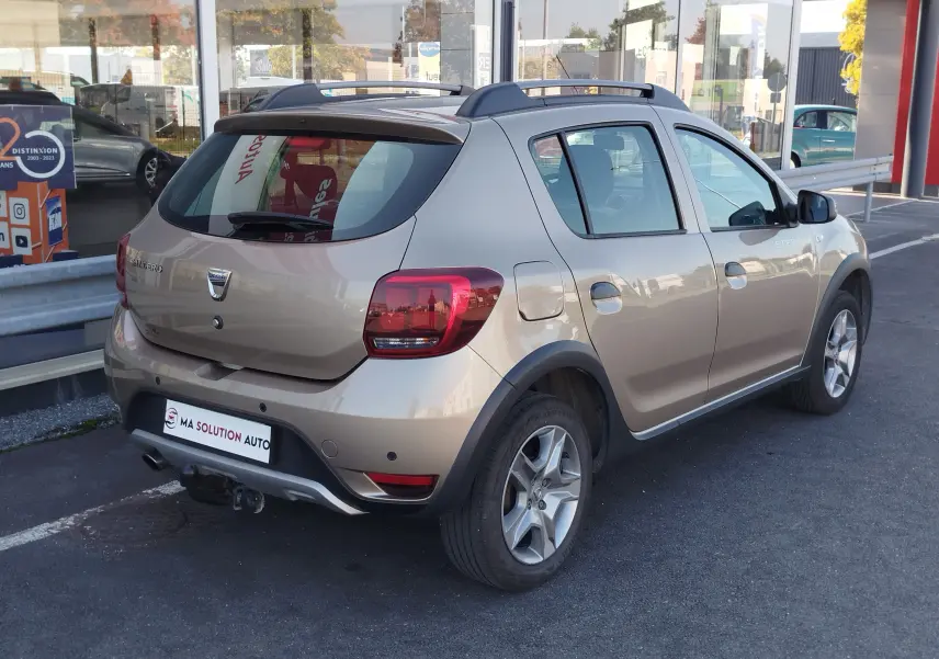 Vue 3/4 arrière droite d'une Dacia Sandero Stepway beige dune 2020 avec barres de toit et protections noires.